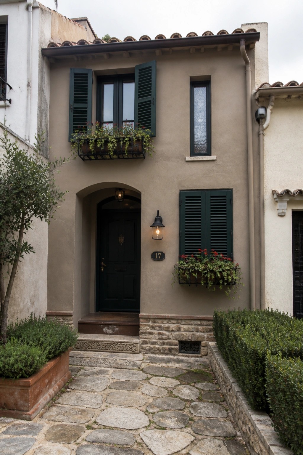 A small two-story house with beige stucco walls, a clay tile roof, dark green shutters on the windows, an arched dark door, a stone base, and a cobblestone path leading to the entrance.