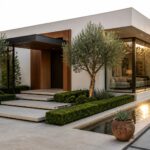 Modern white villa exterior with dark canopy over wooden front door, large glass windows beside the entrance, stone steps, olive trees, and a linear water feature along the paved approach.
