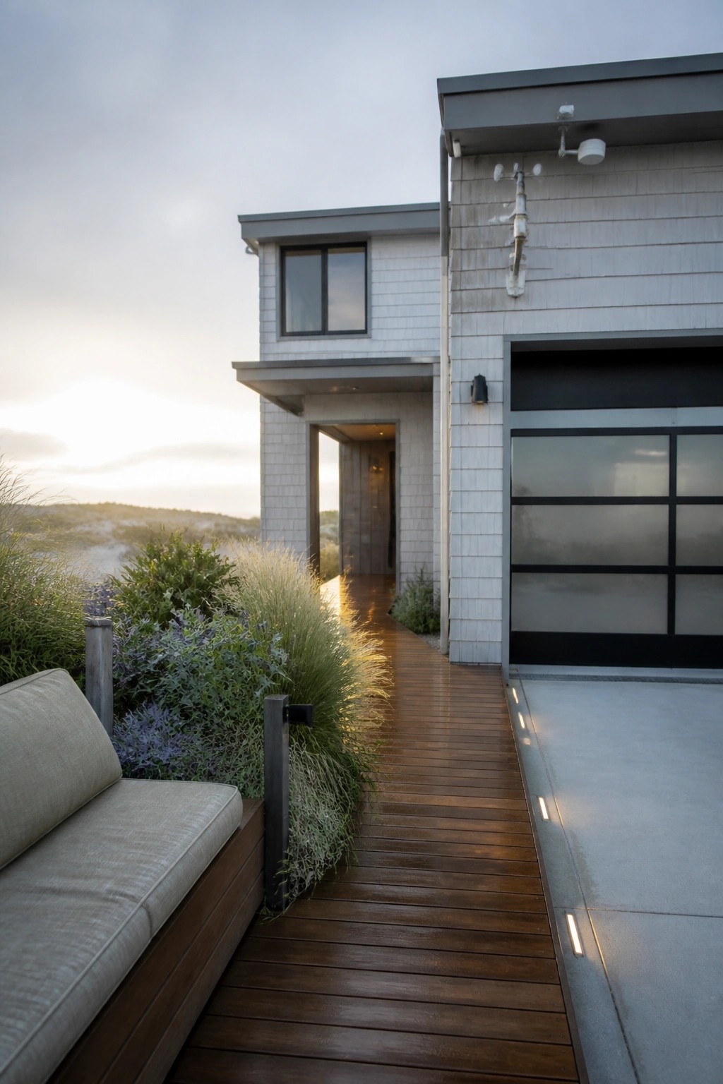 A contemporary house exterior with light shingle siding, a black-framed garage door on the right, and a wooden walkway leading past plants to an open entry door.