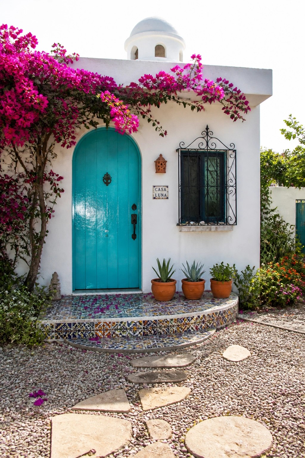 A small white stucco house with a bright turquoise arched door, pink bougainvillea growing over the top, and colorful tiled steps leading to the entrance.