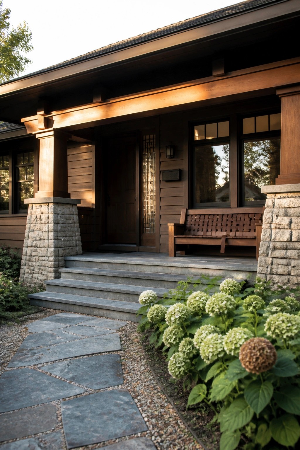 A front porch of a dark wood house with a wide overhanging roof, stone support pillars, gray stone steps, a wooden bench, and white hydrangeas along the path.