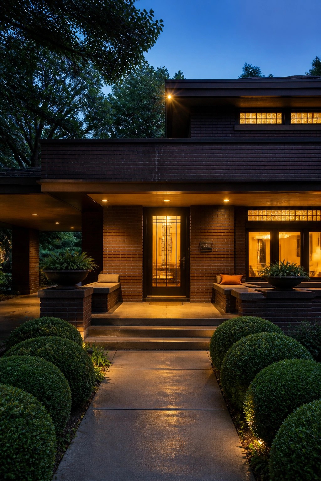 Front view of a brick prairie style house at dusk showing low horizontal rooflines, a central lit entry door, and rounded shrubs along the walkway.