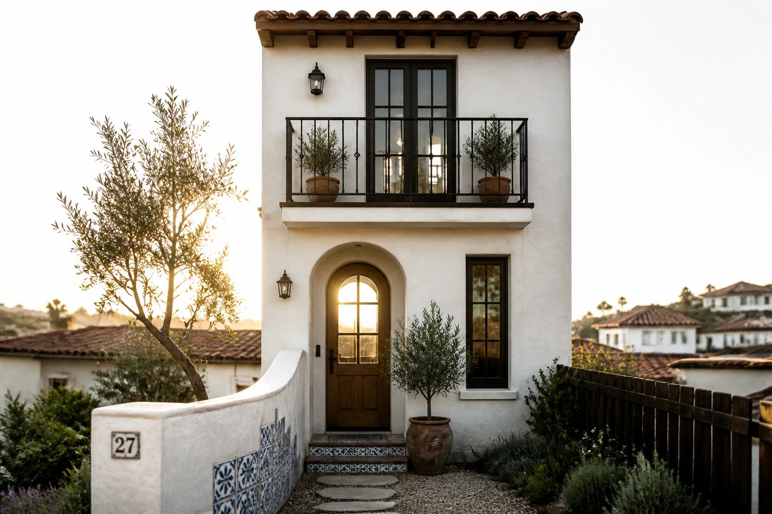 A small two-story Mediterranean home with white stucco walls, a prominent arched wooden front door, and a black metal balcony directly above it.