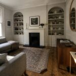 A living room with a white fireplace and mantel, flanked by arched built-in shelves holding books and vases, a gray sofa on a large patterned rug, and a wooden sideboard beneath a window.