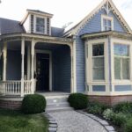 A small blue-gray Victorian house with white trim, a front porch with turned posts, a bay window, gray steps, and a gravel path.
