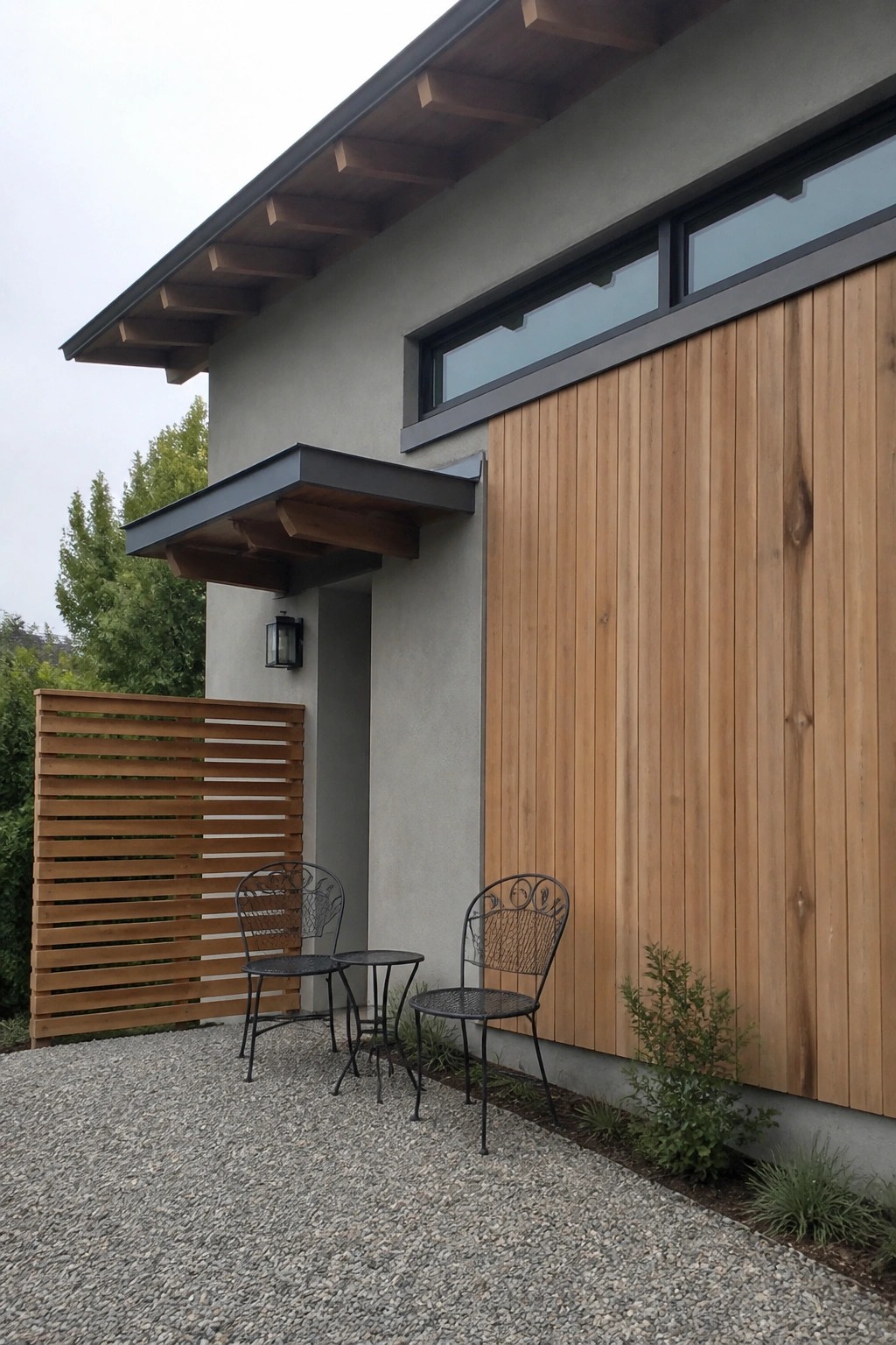 Modern house exterior showing a section of vertical wood siding next to a gray stucco wall, with a small covered entry, two metal chairs on gravel, and a wooden fence.