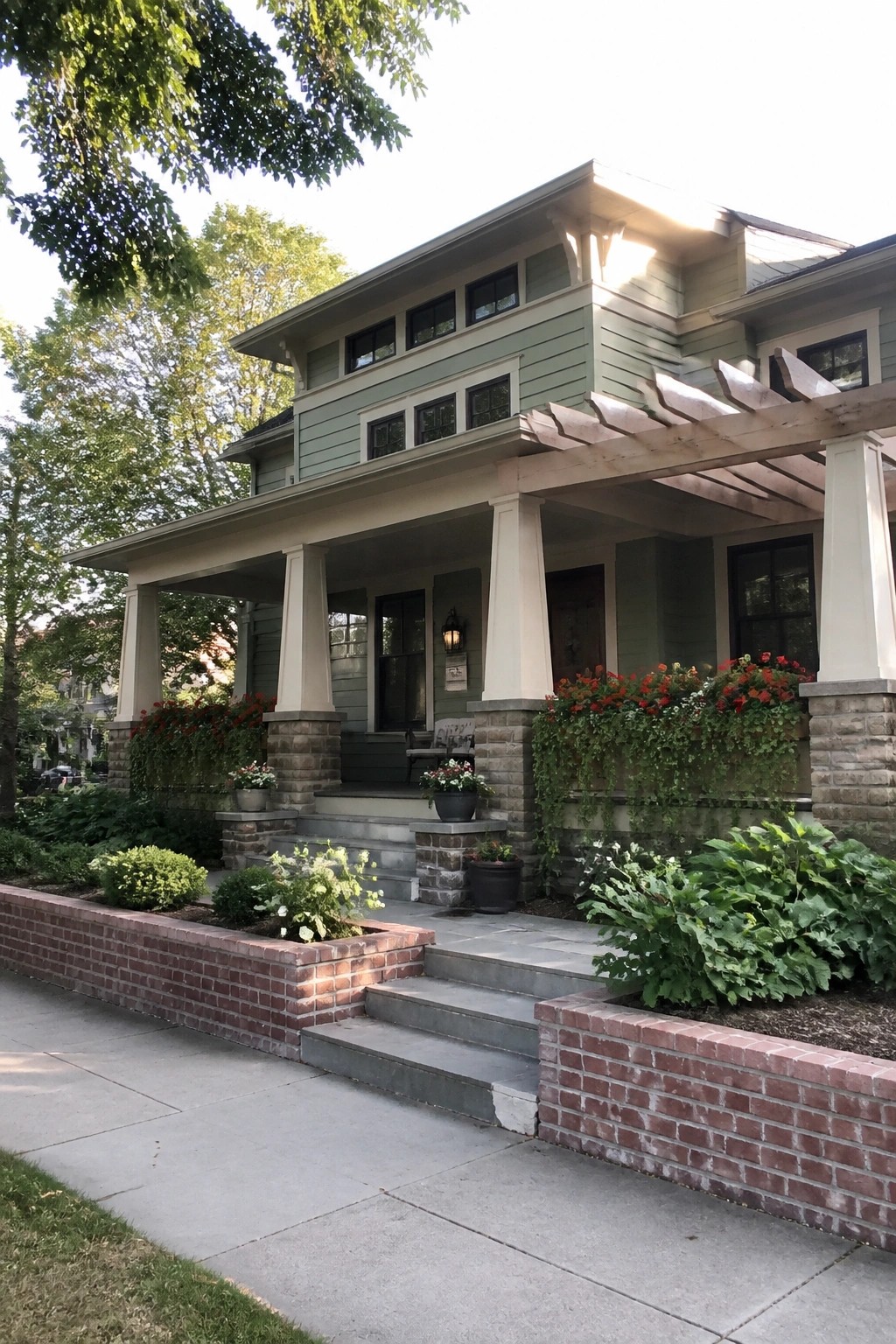 Light green two-story home with porch columns, red flowers, brick walls and stone steps.