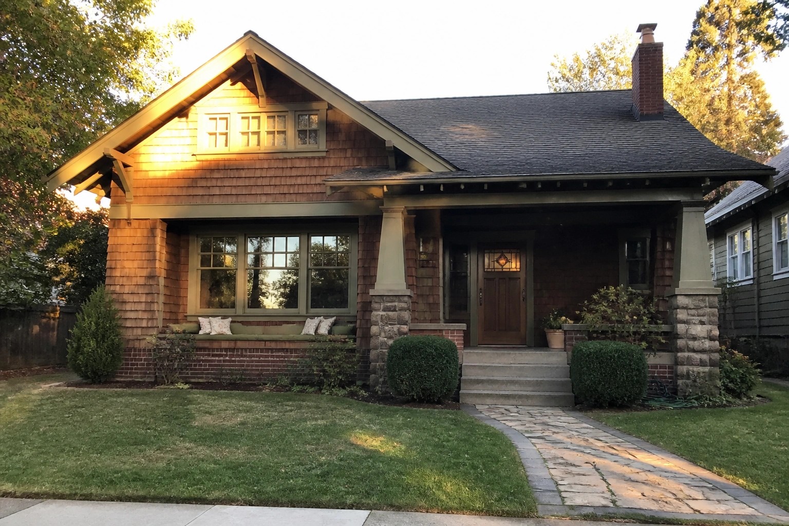 Craftsman bungalow with shingled walls, stone porch pillars, and curved stone path on lawn.