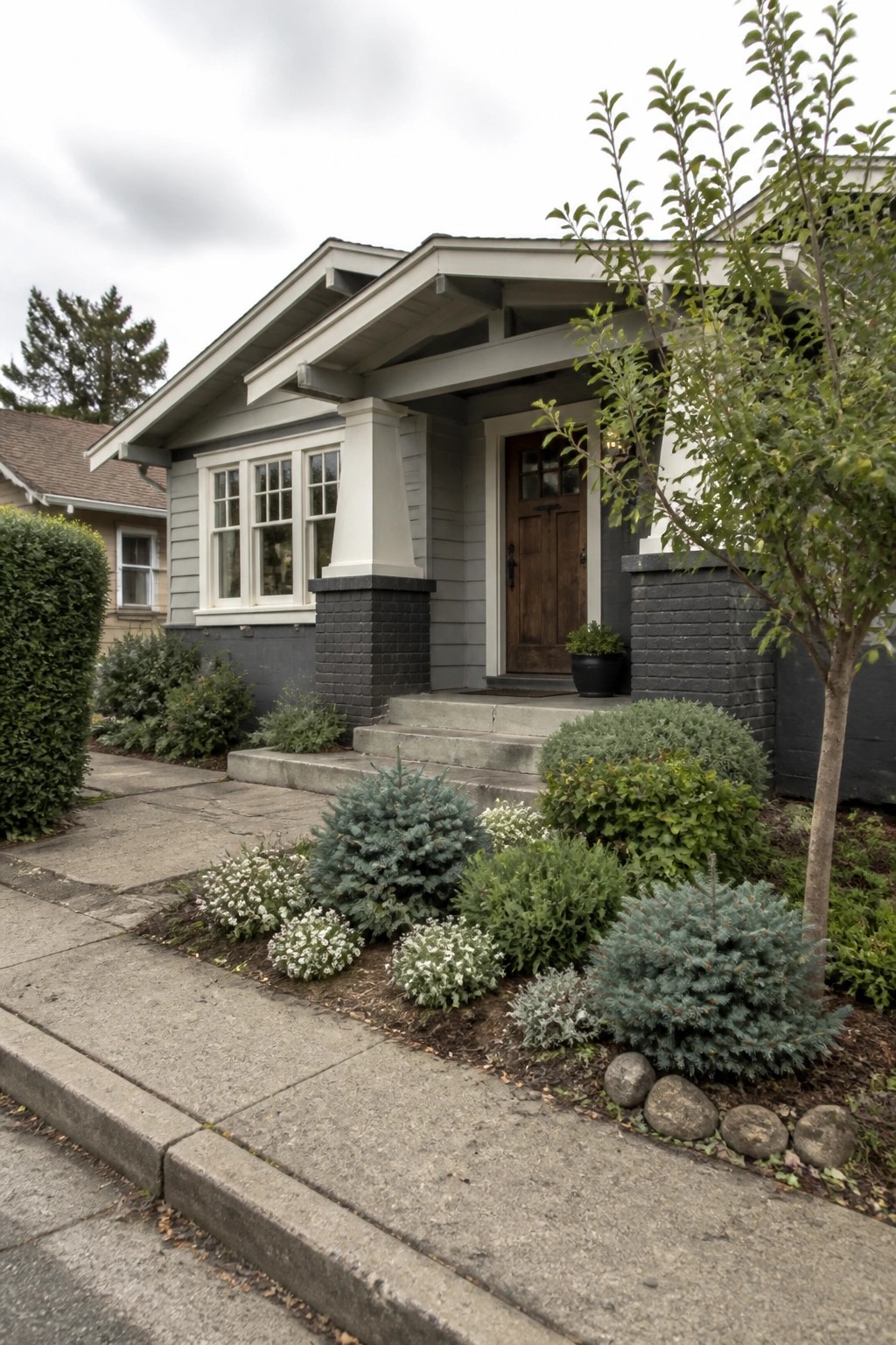 Gray craftsman bungalow with wooden door, white columns, brick pillars, and shrub garden.