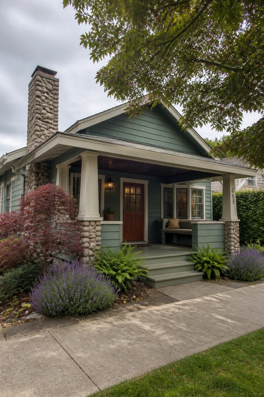 Green Craftsman home with stone chimney, red door, porch, and purple flowers