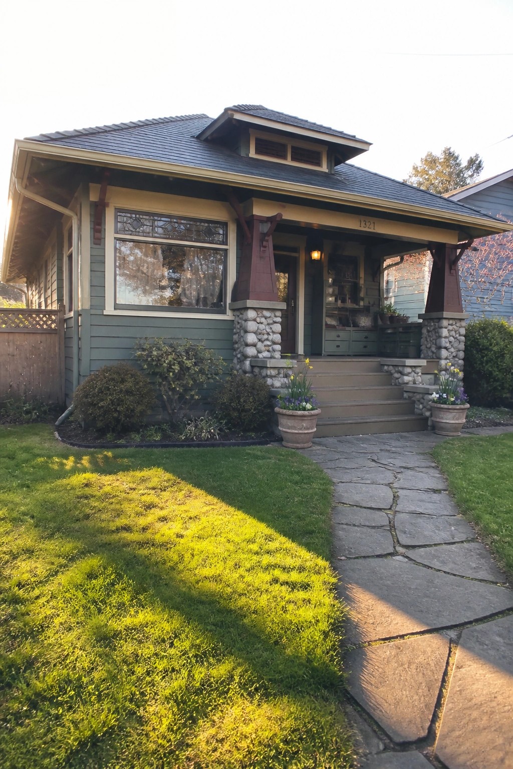 Green Craftsman bungalow with stone porch, steps, potted flowers, and stone path.