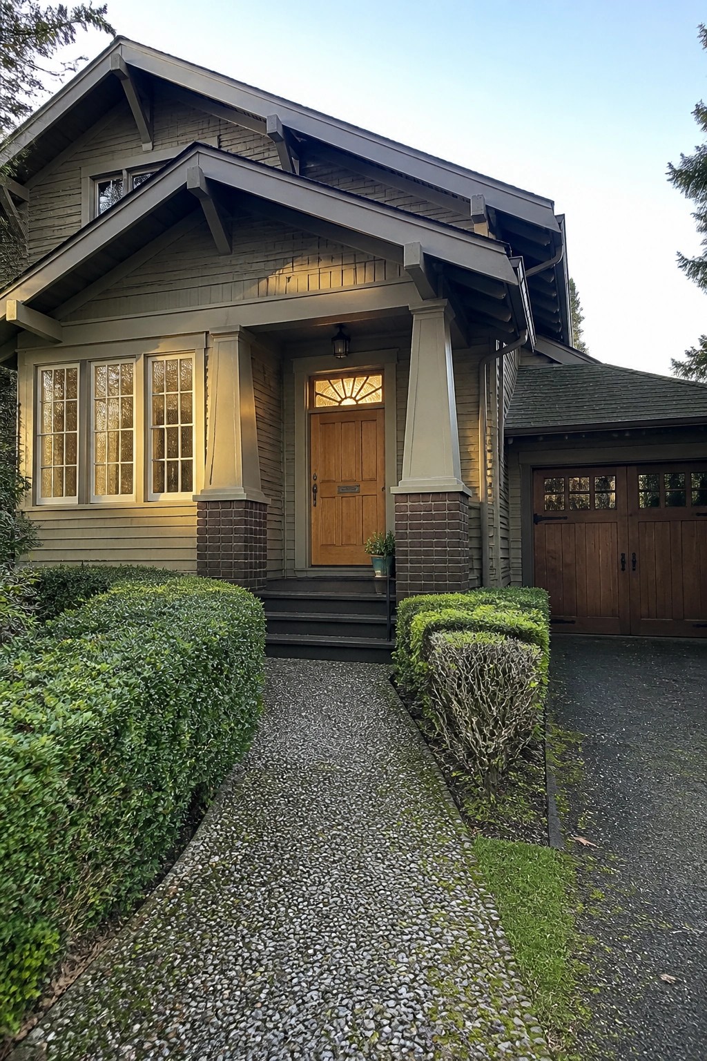 Front view of a Craftsman bungalow showing a gabled porch with tapered columns on brick piers, a centered wooden door, multi-pane windows, and a gravel path with shrubs.