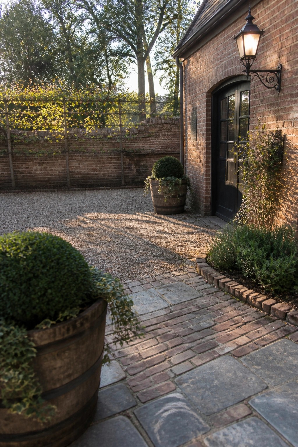 Brick courtyard with cobblestone path, wooden plant barrels, and lit lantern on brick building
