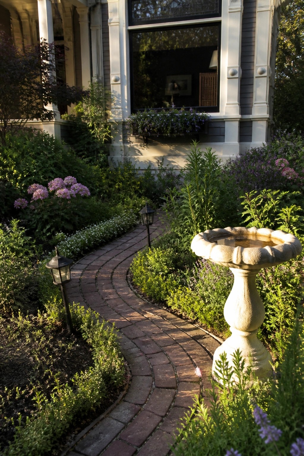 A curving brick pathway winds through dense garden beds with purple flowers and greenery past a stone birdbath toward a gray Victorian house.