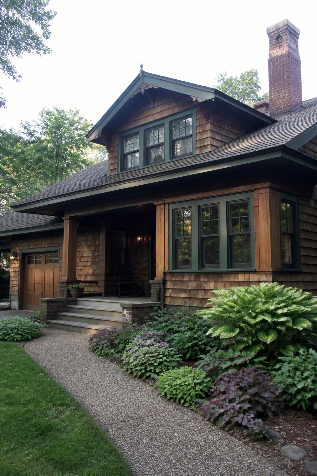 Rustic wooden house with green trim, large windows, brick chimney, and vibrant garden.