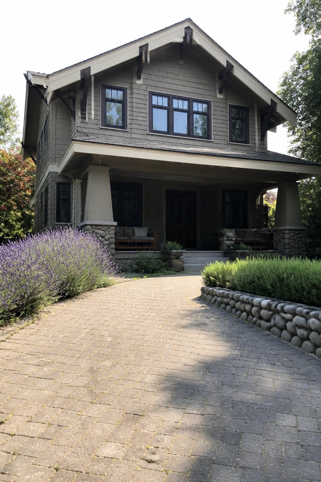Gray Craftsman house with porch, brick driveway, lavender bushes, and green shrubs.