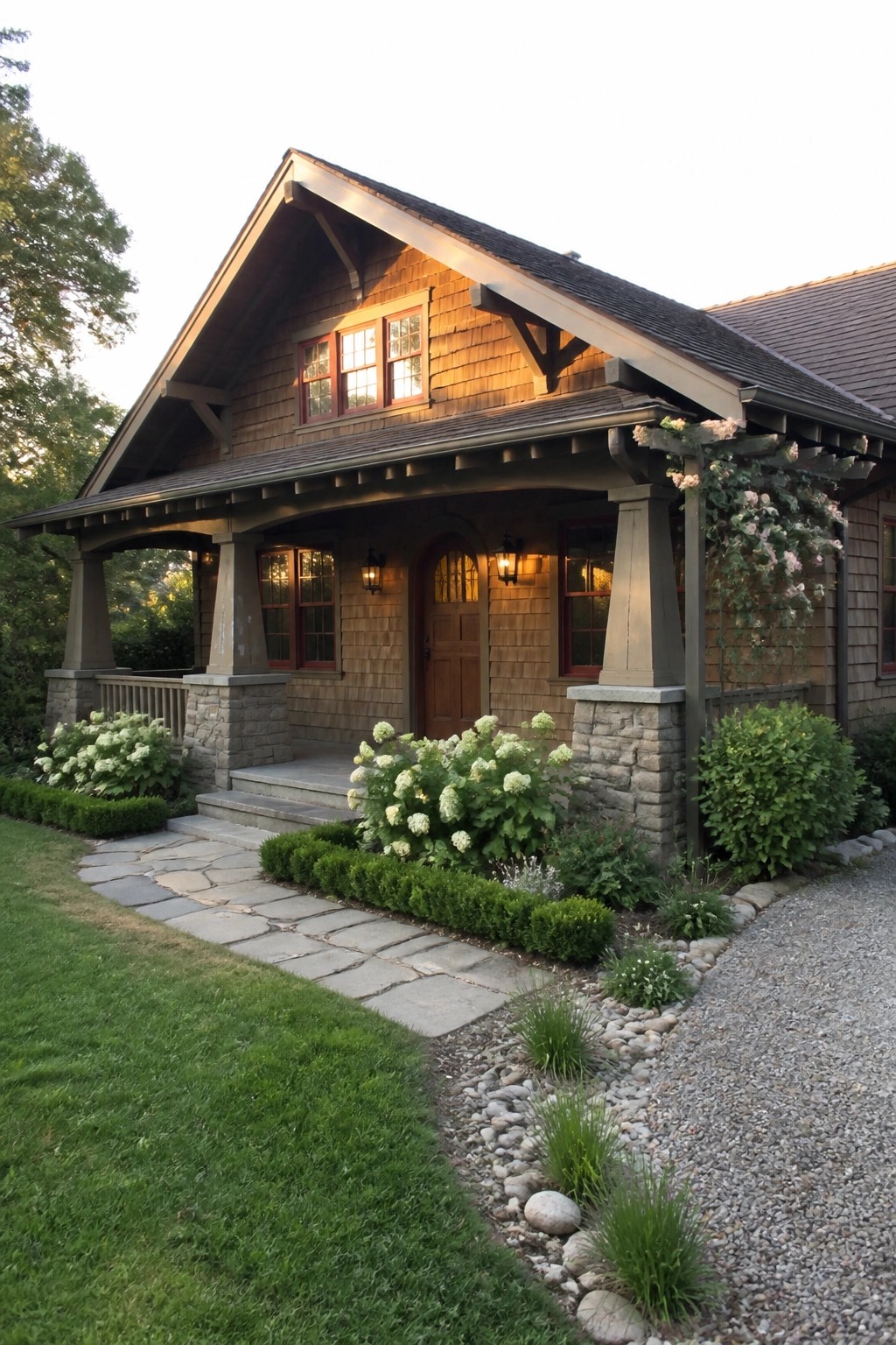 A Craftsman bungalow exterior featuring wood shingle siding, a front porch with tapered stone column bases, an arched entry door, and landscaped beds with white hydrangeas along a stone pathway.