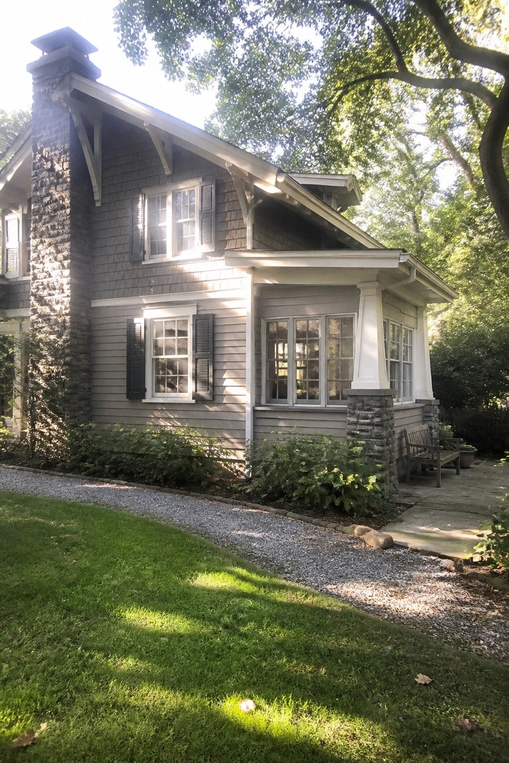 A two-story Craftsman bungalow with gray wood siding, a tall stone chimney, black shutters, a covered porch with a stone base, and a gravel path in front.