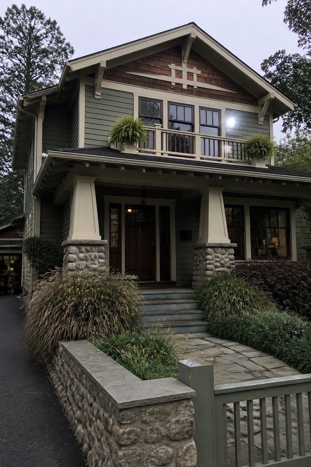 A two-story craftsman house with green siding, a covered front porch supported by tapered pillars on stone bases, and a small upper balcony.