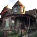 A large Victorian house with a wraparound porch, shingled turret, and stone steps leading to a wooden front door, surrounded by green shrubs and climbing vines.