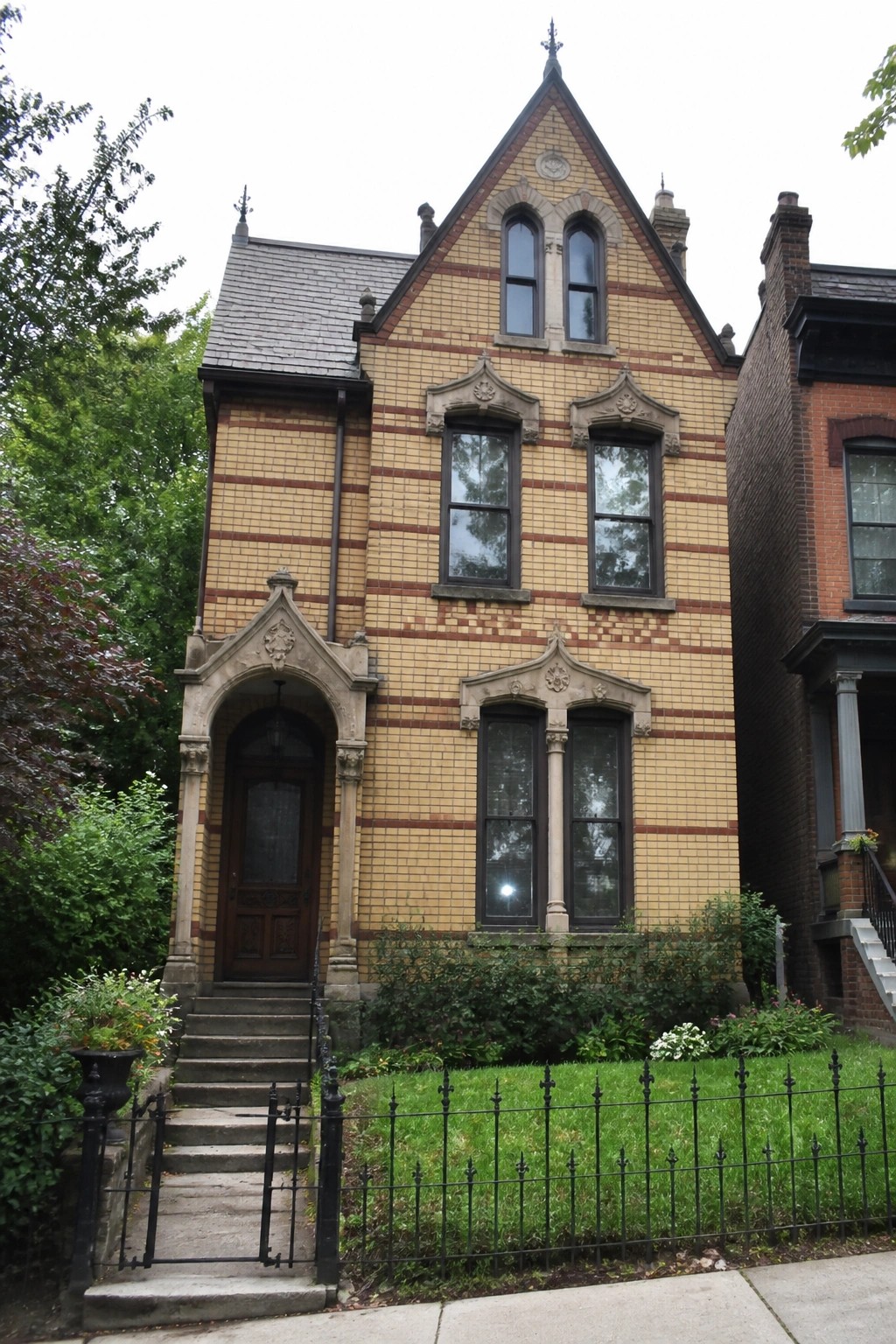 A two-story Victorian house with blue-gray siding, lighter trim, a small upper balcony, and cream double doors with black hardware, shown from the front steps with shrubs and mulch in the foreground.