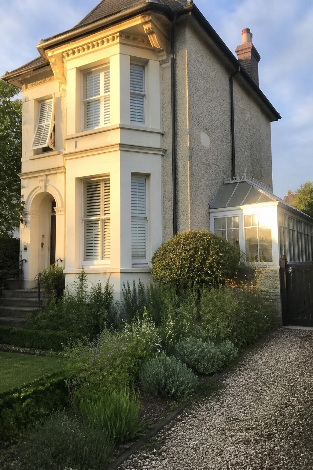 A light green Victorian house with a front porch, brown door, and irregular stone path leading across the lawn to the steps, framed by green hedges with a metal bench on the right.