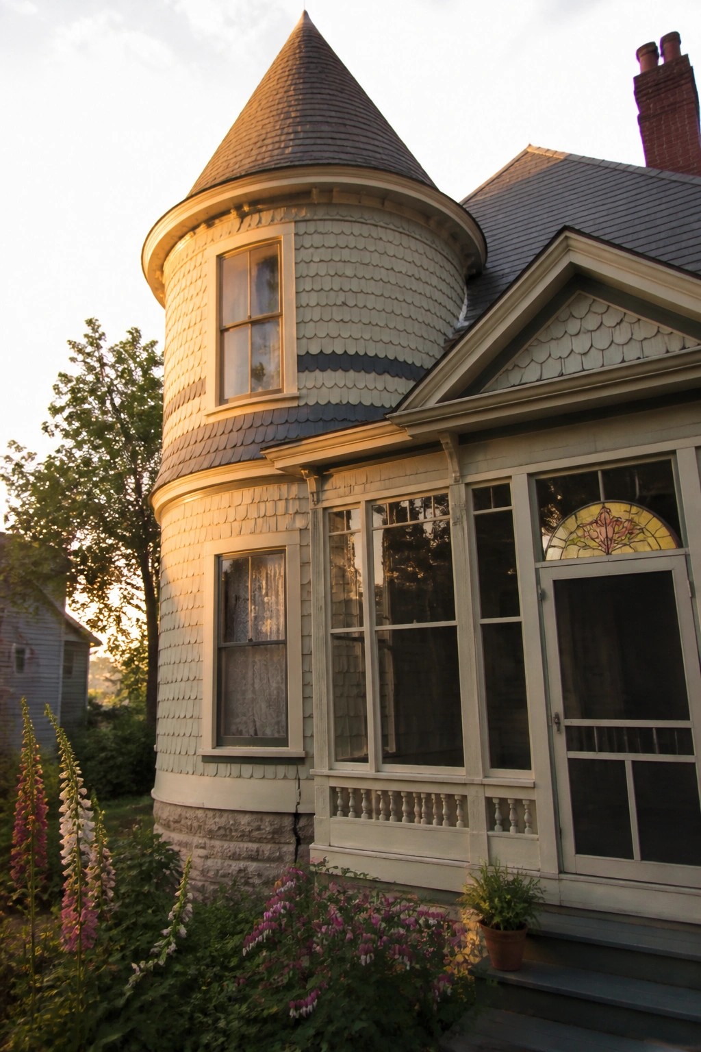 A large gray-shingled Victorian house with a wraparound porch sits beside a sandy beach at sunset.