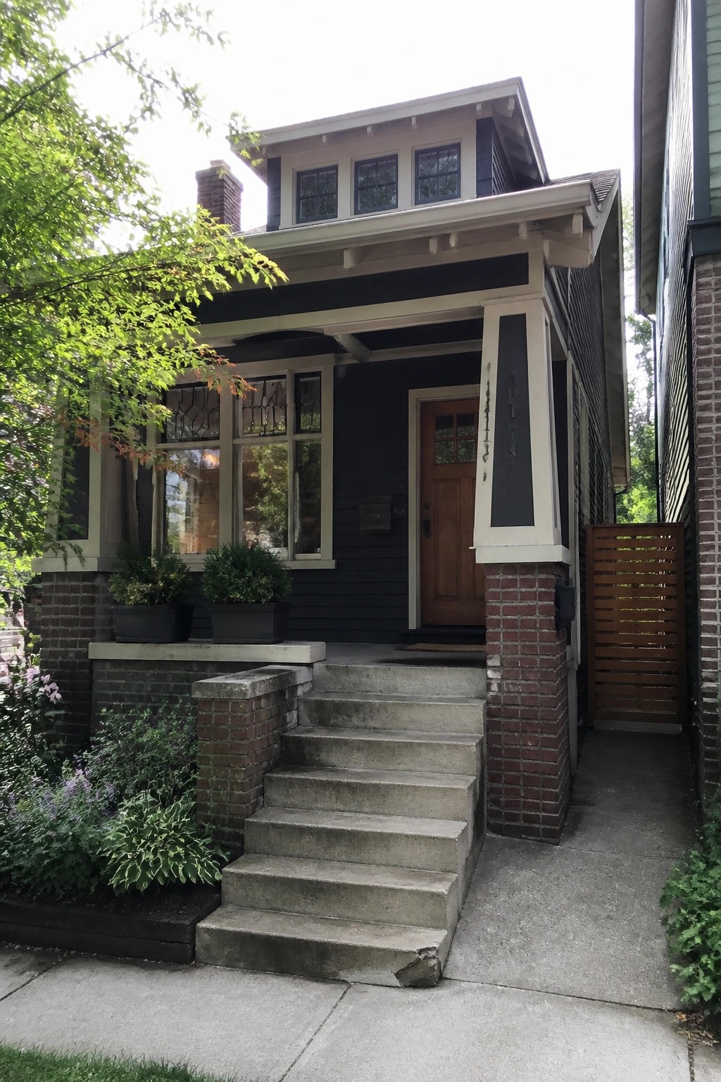 Front view of a dark blue Craftsman house with a porch supported by brick piers and white columns, concrete steps, and a wooden front door.