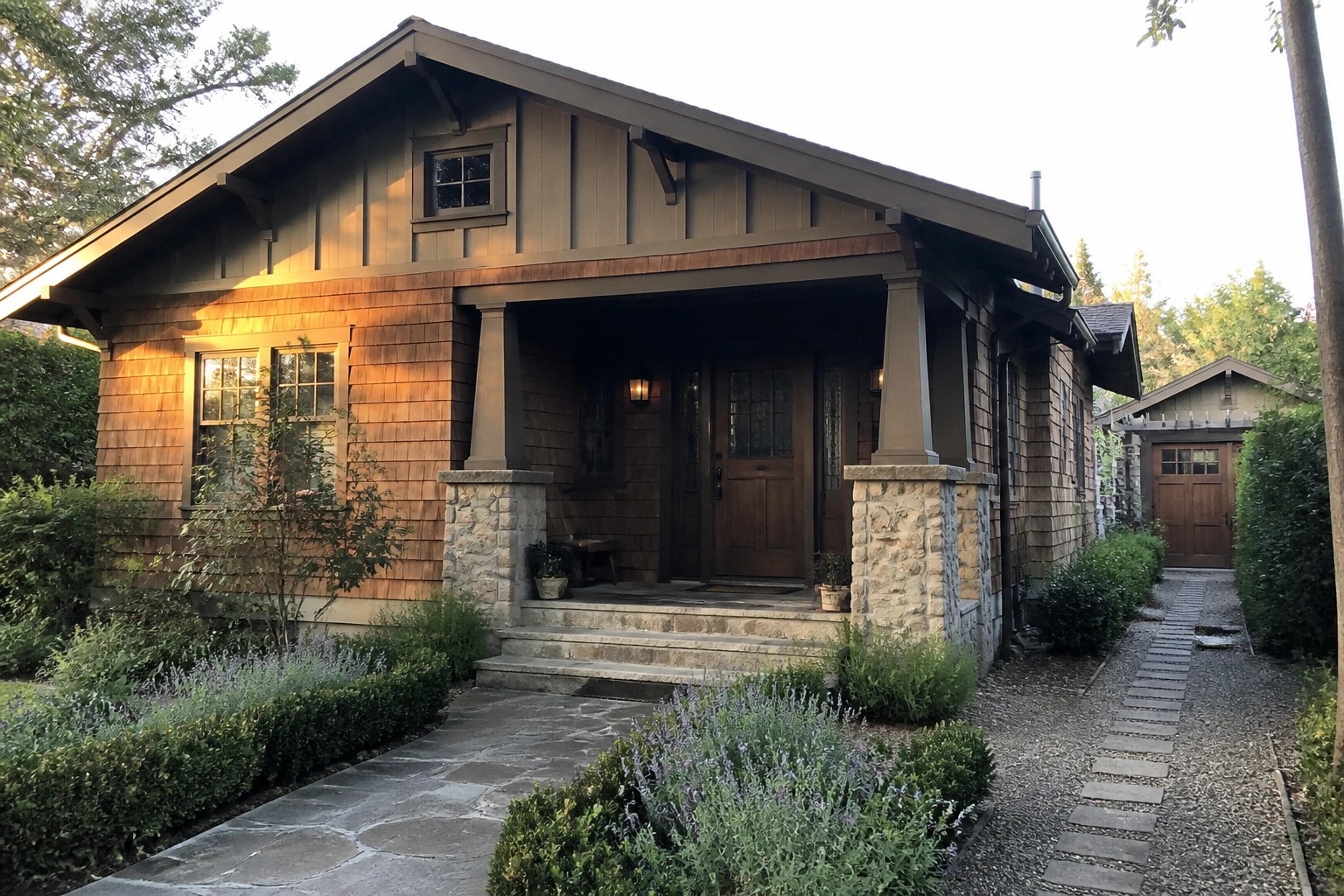 A Craftsman style house exterior featuring a front porch with stone piers, wood siding, a wooden door, and a stone pathway bordered by shrubs and lavender.