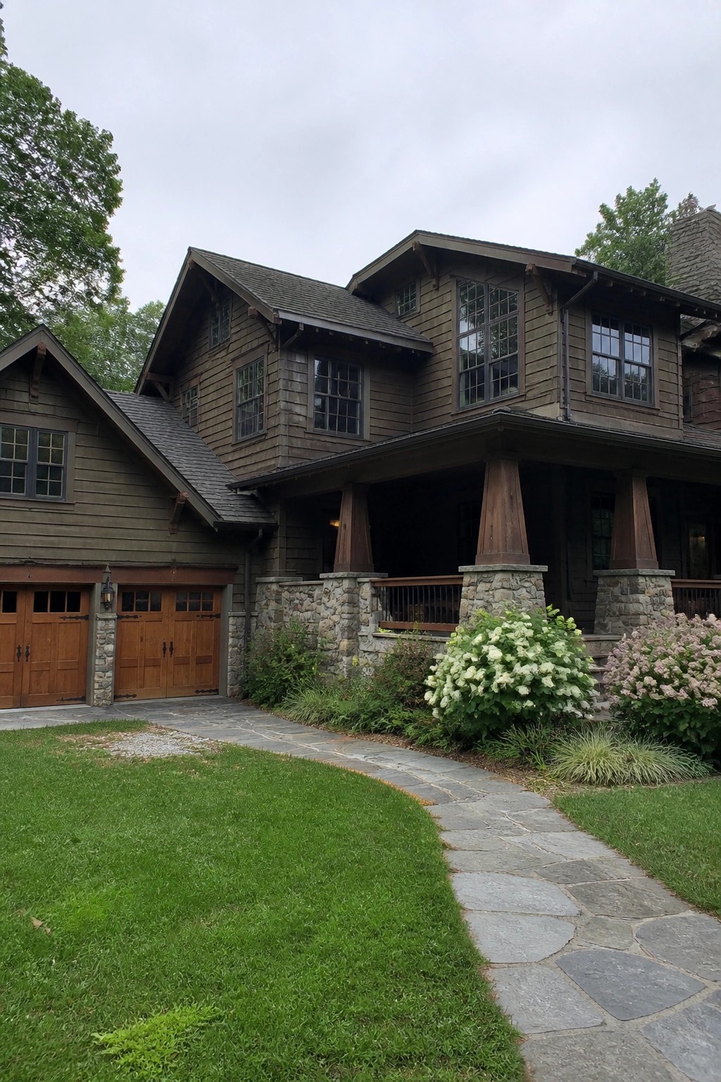 A Craftsman house with brown wood siding, stone foundation and porch piers, wooden garage doors, and a stone pathway leading to the entrance.