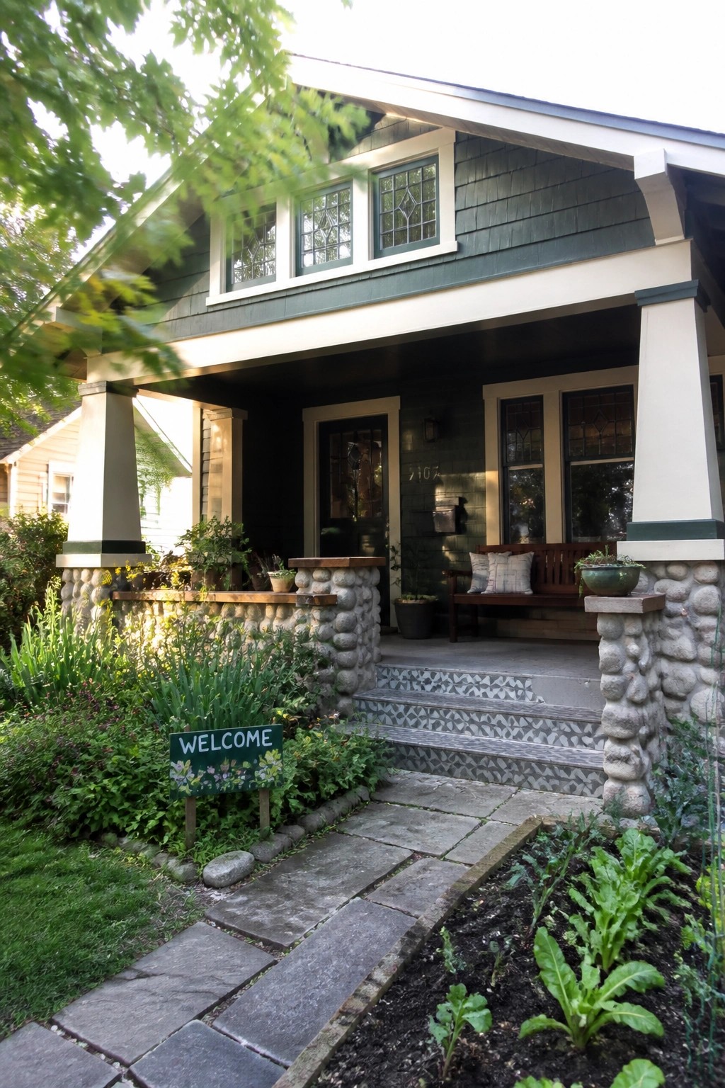 Stone pathway to green Craftsman house porch with welcome sign and garden.
