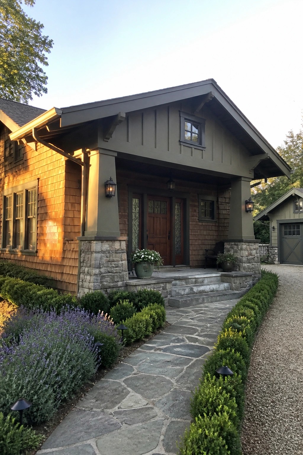 A Craftsman style house exterior featuring a front porch with stone piers, wood siding, a wooden door, and a stone pathway bordered by shrubs and lavender.