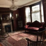 Victorian parlor with red velvet sofa, crystal chandelier, fireplace, and sunlit rug.