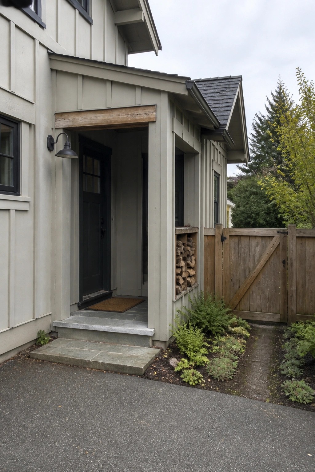 A light gray Craftsman house with a dark front door under a covered entry, stone steps, and a built-in firewood rack beside a wooden gate.