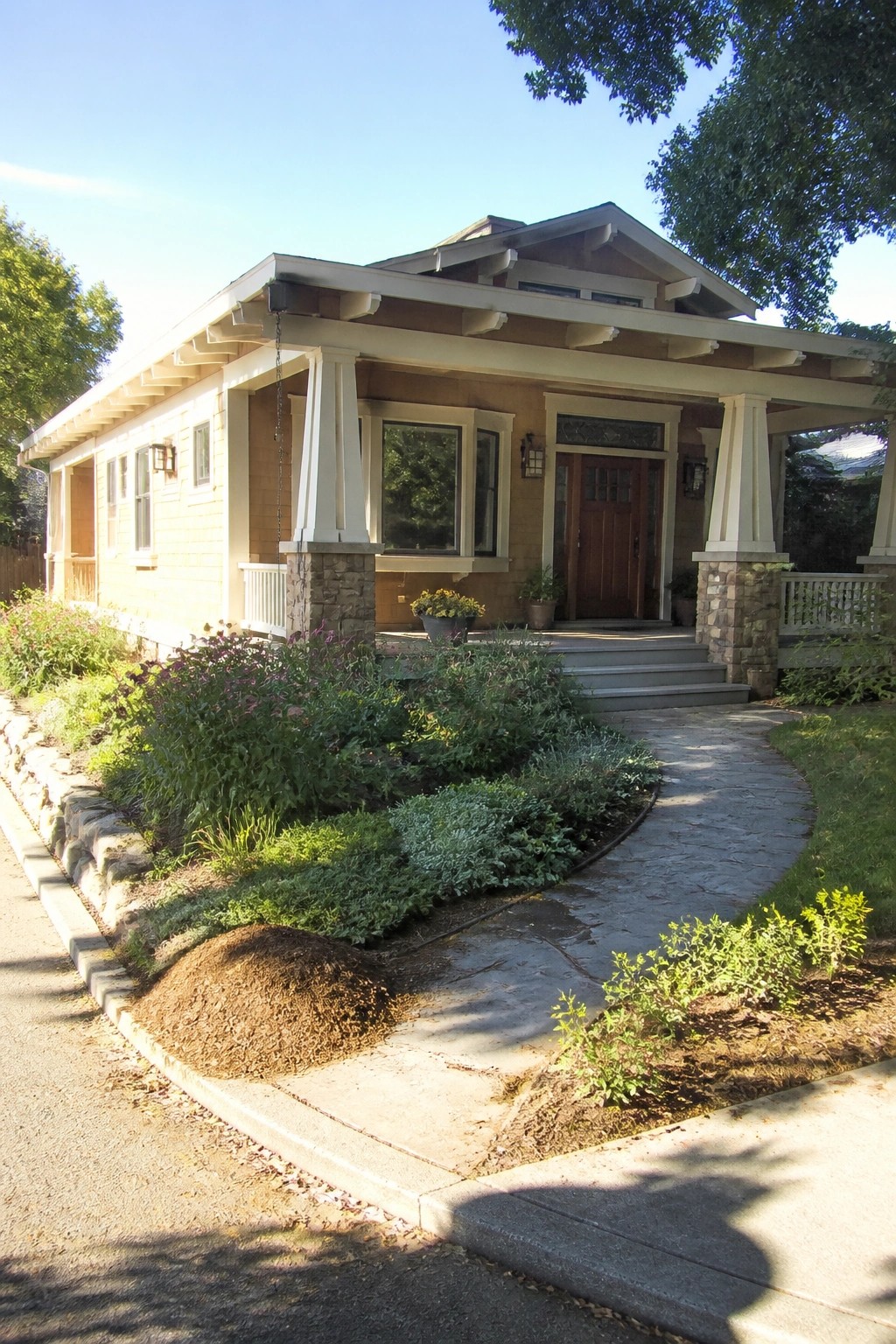 Craftsman bungalow with beige siding, stone porch pillars, wooden door, curving path, and garden.