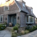 A coastal cottage exterior with weathered gray shingle siding, a blue arched front door, dormer windows, and hydrangeas along a stone pathway.