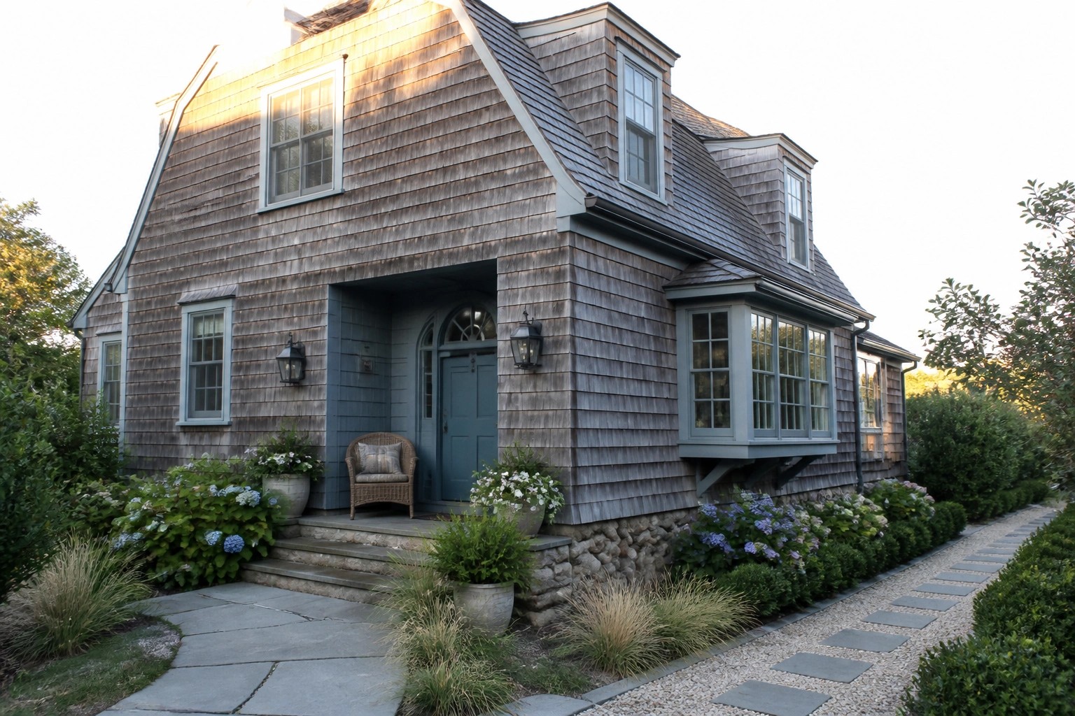 A coastal cottage exterior with weathered gray shingle siding, a blue arched front door, dormer windows, and hydrangeas along a stone pathway.