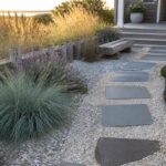 A gravel garden path with irregular gray stepping stones winds past a low stone wall, tall native grasses, and clumps of purple flowering plants.