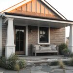 A small gray shingled beach cottage with a gabled roof, white columns, a wooden front porch bench with striped cushions, and stone steps leading to a paver path.