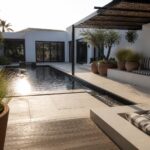 A modern courtyard with a long rectangular pool, white walls, potted olive tree and grasses, built-in concrete benches with striped cushions, and a dark wooden pergola overhead.
