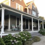 A gray shingled coastal house with a white wraparound porch that includes a hanging swing, two Adirondack chairs, and blue hydrangeas along the front steps.