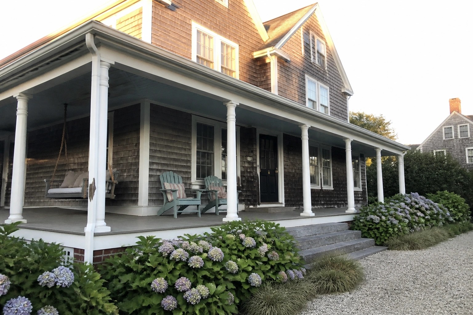 A gray shingled coastal house with a white wraparound porch that includes a hanging swing, two Adirondack chairs, and blue hydrangeas along the front steps.