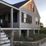 A two-story shingled house elevated on concrete piers with a front porch, wooden stairs, and a low stone retaining wall along the side.