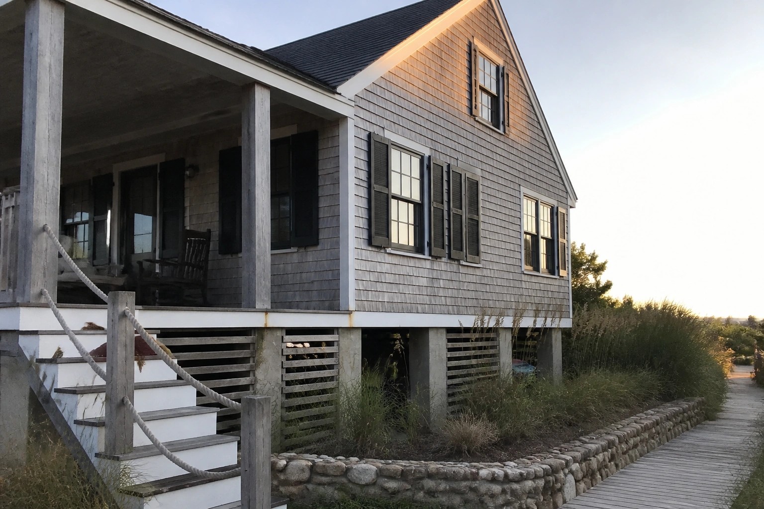 A two-story shingled house elevated on concrete piers with a front porch, wooden stairs, and a low stone retaining wall along the side.