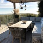 A long wooden dining table with woven chairs on one side and a light blue cushioned bench on the other sits on a gray deck under a wooden pergola, with an outdoor kitchen visible in the background and dunes beyond a glass railing.