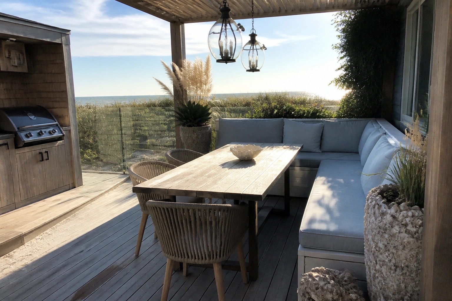 A long wooden dining table with woven chairs on one side and a light blue cushioned bench on the other sits on a gray deck under a wooden pergola, with an outdoor kitchen visible in the background and dunes beyond a glass railing.