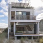 Modern white two-story beach house with open glass doors, balcony, and deck